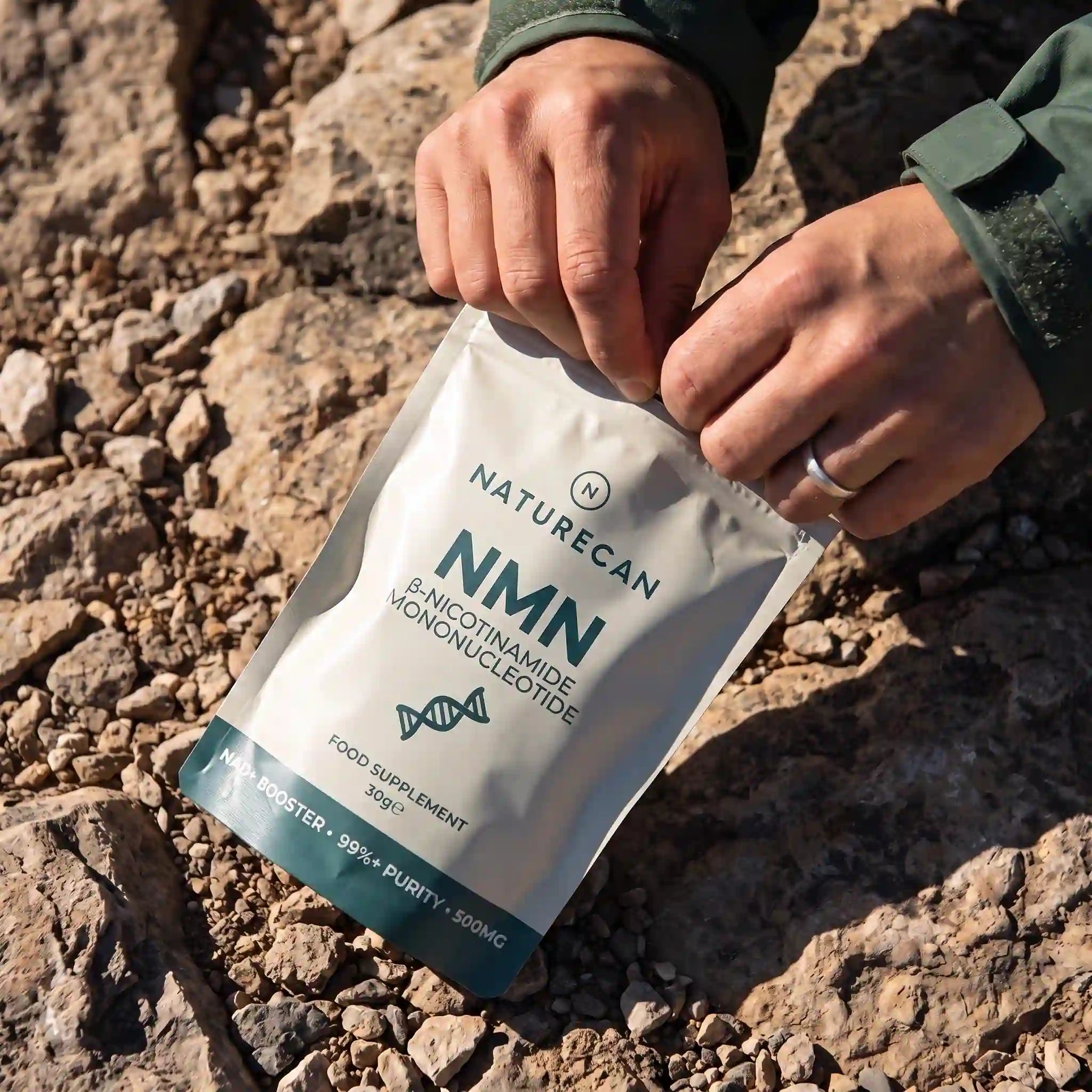 A person's hands are shown opening a Naturecan NMN food supplement pouch on a rocky, outdoor gravel surface.