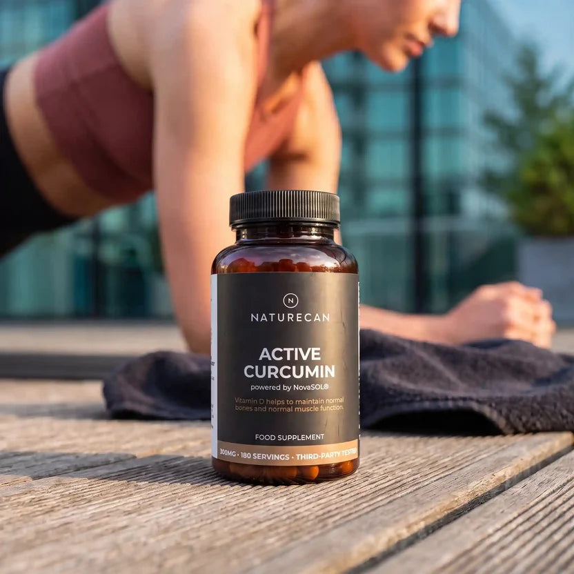 A bottle of Naturecan Active Curcumin food supplement placed on a wooden table in an outdoor setting, with a woman in athletic wear blurred in the background.
